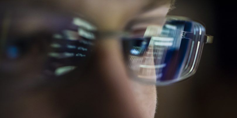 Portait of young hacker with computer screen reflecting in the man's glasses.